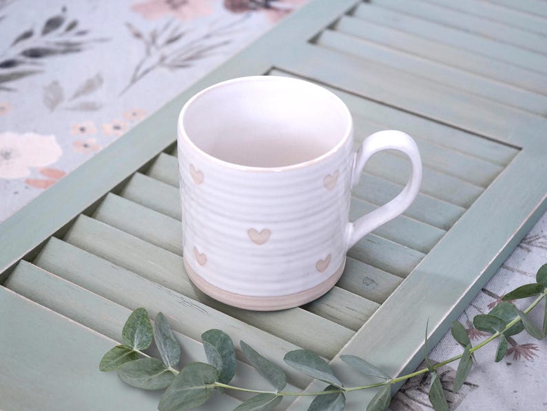 May include: A white ceramic mug featuring a ribbed texture and small, light-colored heart patterns scattered across the exterior. The mug has a curved handle and a smooth, unglazed base, showcasing a simple design with a clean, rounded silhouette.