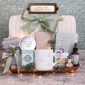 Op de afbeelding: Een geschenkset met het label Healing Escape Box, bestaande uit een cr&egrave;me kabelgebreide plaid van 124 bij 155 cm, een mok met madeliefjespatroon, een geschulpt schaaltje, pepermuntpapierzeep, zandkoekjes, honing, een dipper en linnenspray.