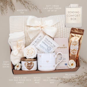 May include: A gift box containing a 49 by 61 inch cream lace knit throw with a satin bow, cream socks, Tea with Tae tea bags, a Hug in a Jar soy candle, a heart-patterned mug, a Moonstruck milk chocolate bar, and a gold pouch of muscle soak.