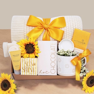 May include: A gift box containing a cream-colored knit blanket with a yellow ribbon, a sunflower, a tube of Deep Steep body lotion, a Sun Shine bath bomb, a box of hot chocolate, a white mug with a small dish, and a Moonstruck chocolate bar.