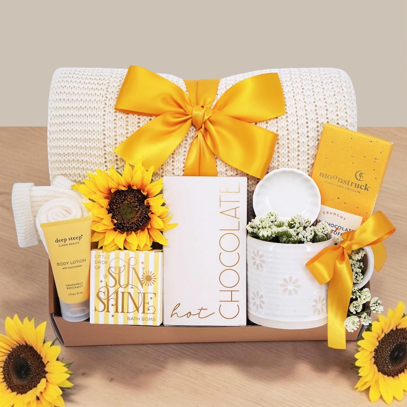 May include: A gift box containing a cream-colored knit blanket with a yellow ribbon, a sunflower, a tube of Deep Steep body lotion, a Sun Shine bath bomb, a box of hot chocolate, a white mug with a small dish, and a Moonstruck chocolate bar.