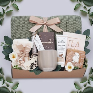 K&ouml;nnte beinhalten: Ein Geschenk-Korb mit Pflegeprodukten. Er enth&auml;lt eine gr&uuml;ne Strickdecke, eine beige Schleife, eine Kerze, Handcreme, Tee-Proben, einen Vogel-Ornament aus Holz und Beinw&auml;rmer. Der Korb ist mit Gr&uuml;n und wei&szlig;en Blumen dekoriert.