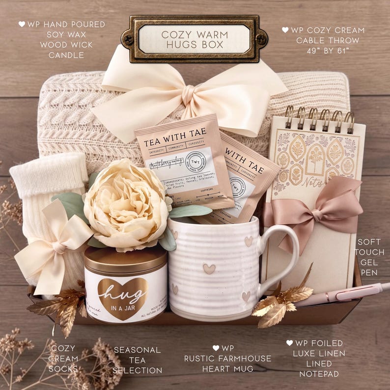 Op de afbeelding: Een geschenkdoos met het label COZY WARM HUGS BOX met een cr&egrave;mekleurige kabelgebreide deken van 124,46 bij 154,94 cm, een sojawas kaars met houten lont, twee Tea With Tae theezakjes, een cr&egrave;mekleurige mok met hartjes, een notitieblok en sokken.