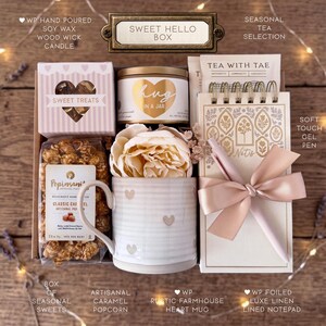 Op de afbeelding: Een geschenkdoos met het label Sweet Hello Box bevat een handgegoten sojawas kaars, een zak karamel popcorn, een keramische mok met hartjespatroon, een kunstpioenroos, een selectie thee, een notitieblok en een gelpen met een strik.