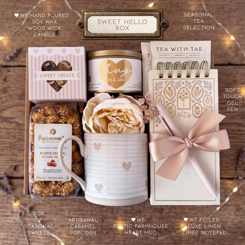 Op de afbeelding: Een geschenkdoos met het label Sweet Hello Box bevat een handgegoten sojawas kaars, een zak karamel popcorn, een keramische mok met hartjespatroon, een kunstpioenroos, een selectie thee, een notitieblok en een gelpen met een strik.