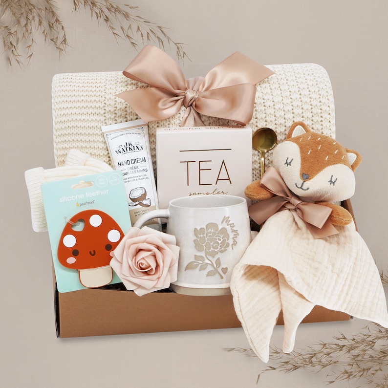 May include: A gift box containing a cream-colored knit blanket with a large satin bow, a tube of J.R. Watkins hand cream, a tea sampler box, a ceramic mug with a floral design and the word mama, a mushroom silicone teether, and a fox plush toy.