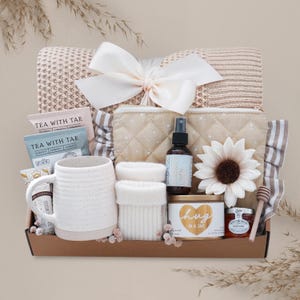 May include: A gift basket filled with a cream-colored blanket, a white mug, tea packets, a spray bottle, a candle, honey, and a decorative flower. The basket is tied with a large white bow.