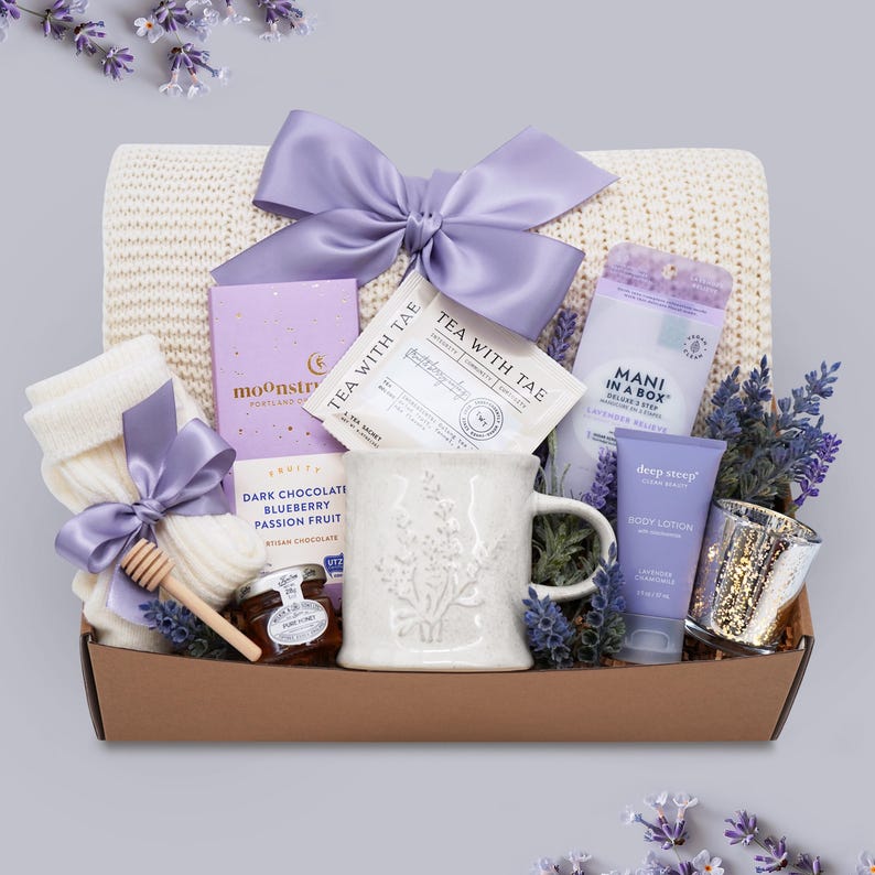May include: A gift basket filled with self-care items. It includes a white mug with floral detailing, tea sachets, a blanket, socks, chocolate, honey, body lotion, and a candle holder. The items are arranged in a brown box and tied with a lavender bow.