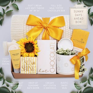 May include: A gift box with a soft cream lace knit throw (49" x 61"), a sunflower, and a yellow bow. Includes a chocolate bar, lotion, bath bomb, hot cocoa, a daisy mug, and a trinket dish. The box is labeled "Sunny Days Ahead".