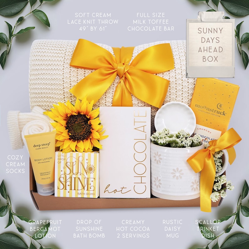 May include: A gift box with a soft cream lace knit throw (49" x 61"), a sunflower, and a yellow bow. Includes a chocolate bar, lotion, bath bomb, hot cocoa, a daisy mug, and a trinket dish. The box is labeled "Sunny Days Ahead".