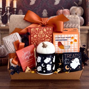 May include: A gift basket containing a beige knit blanket, a red book with gold pumpkin illustrations, a box of Spicy Pumpkin black tea, a black mug with white ghost patterns topped with a white pumpkin, and a box of candy corn undereye masks.