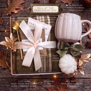 Puede incluir: Una caja de regalo etiquetada como Fall at the Farm Stand Box que contiene una bufanda de cuadros en verde musgo y crema, una cuchara dorada, paquetes de t&eacute;, una taza de cer&aacute;mica blanca moteada, una cinta verde oscuro y una vela de calabaza.