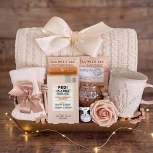 May include: A gift basket containing a cream-colored cable knit blanket with a large satin bow, a pair of white knit socks with a satin bow, a box of Voesh Pedi in a Box Honey Oatmilk, two Tea with Tae packets, a honey dipper, a jar of honey, and a mug.