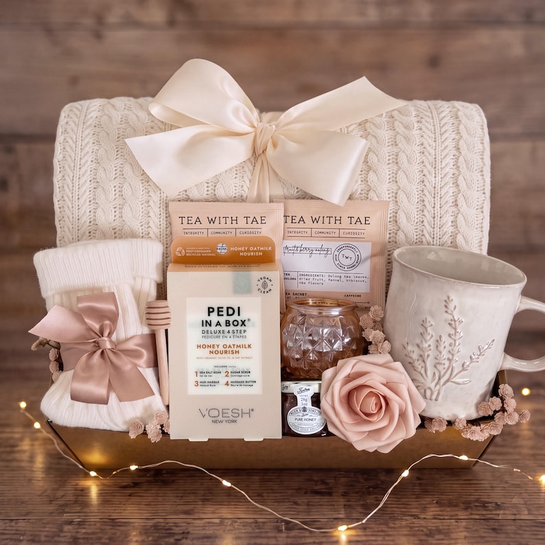 May include: A gift basket containing a cream-colored cable knit blanket with a large satin bow, a pair of white knit socks with a satin bow, a box of Voesh Pedi in a Box Honey Oatmilk, two Tea with Tae packets, a honey dipper, a jar of honey, and a mug.
