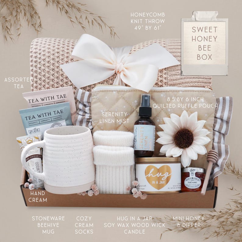 May include: A gift box with a honeycomb knit throw (49 x 61 inches), assorted tea, a stoneware mug, cozy cream socks, a soy wax candle, a mini honey jar, and a quilted ruffle pouch. The box is tied with a large white bow and includes a Serenity Linen Mist.