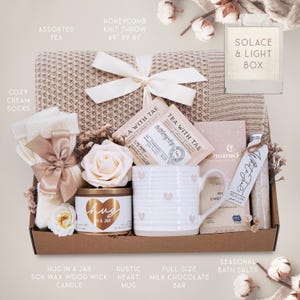 May include: A gift box containing a beige honeycomb knit throw measuring 49 by 61 inches, cream socks with a satin bow, a Tea with Tae sachet, a Moonstruck milk chocolate bar, a white mug with heart details, a Hug in a Jar candle, and bath salts.