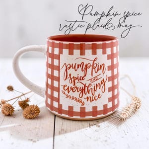 May include: A ceramic mug with a white and orange plaid pattern and a speckled finish. The front features the text "pumpkin spice AND everything nice" in orange cursive script. The interior of the mug is a solid orange color.