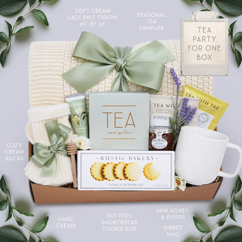 May include: A gift basket with a soft cream lace knit throw (49" by 61"), tea sampler, cozy cream socks, vanilla shortbread cookies, hand cream, mini honey, and a ribbed mug. The basket is tied with a sage green bow.