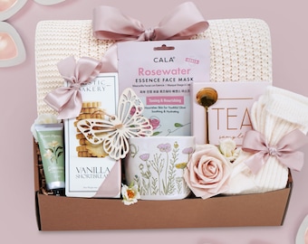 Puede incluir: Una cesta de regalo llena de artículos de cuidado personal, que incluyen una mascarilla facial, galletas de mantequilla de vainilla, una taza floral y una rosa. La cesta está atada con lazos rosas e incluye una manta.