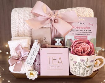May include: A gift set featuring a cream cable-knit blanket tied with a large pink ribbon, a Cala Rosewater face mask, a pink tea sampler box, a mug with floral illustrations, a small jar of honey with a dipper, and a bottle labeled Vanille.