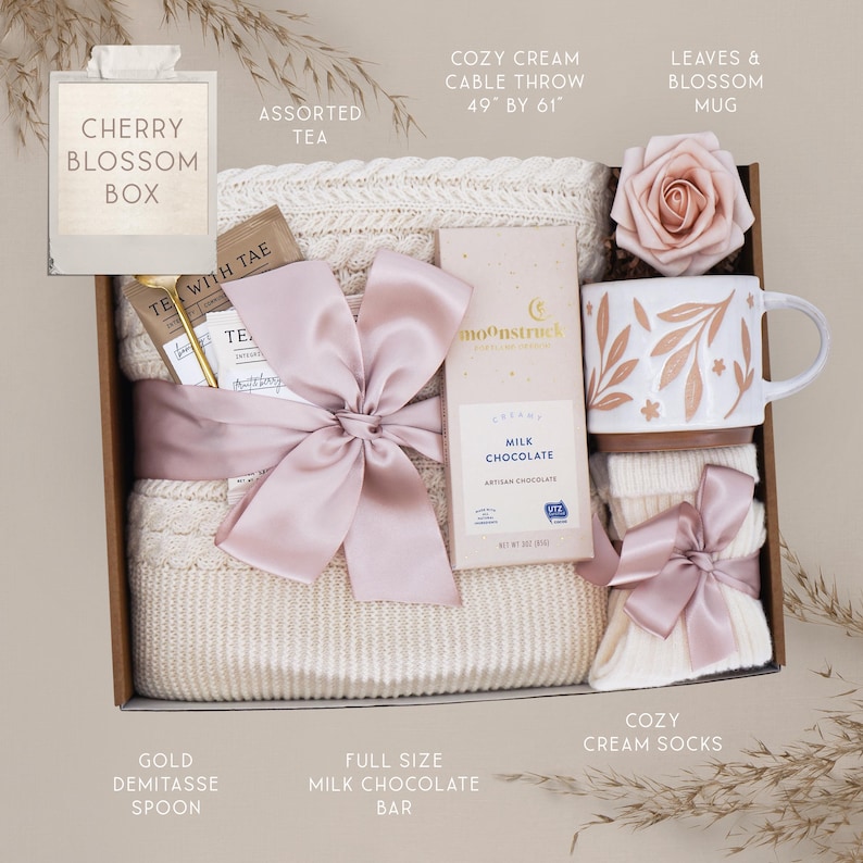 May include: A gift box with a cream-colored cable knit throw (49" x 61"), assorted tea, a milk chocolate bar, a mug with leaf and blossom design, cream socks, and a pink bow. The box is labeled "Cherry Blossom Box".