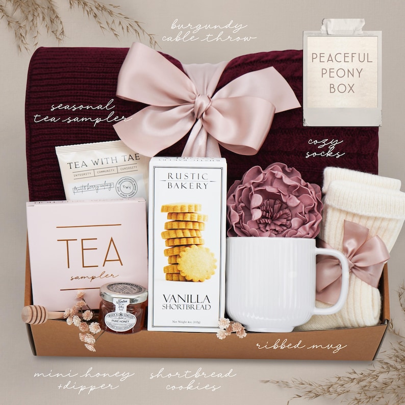 May include: A gift box filled with a burgundy cable throw, cozy socks, a ribbed mug, tea samples, vanilla shortbread cookies, and mini honey with a dipper. The box is tied with a large pink bow and includes a decorative peony.