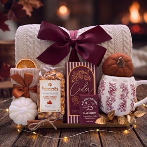 May include: A gift set featuring a cream-colored cable knit blanket with a large burgundy bow, a white pumpkin-shaped mug with floral patterns, a bag of Popinsanity caramel popcorn, a box of cider, a blackberry goat milk soap, and a white pumpkin candle.