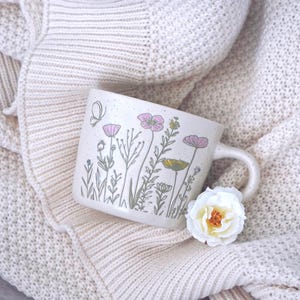 May include: A speckled off-white ceramic mug featuring a delicate botanical illustration of wildflowers in shades of pink, yellow, and green, accompanied by a small butterfly. A single white flower with a yellow center rests near the handle.