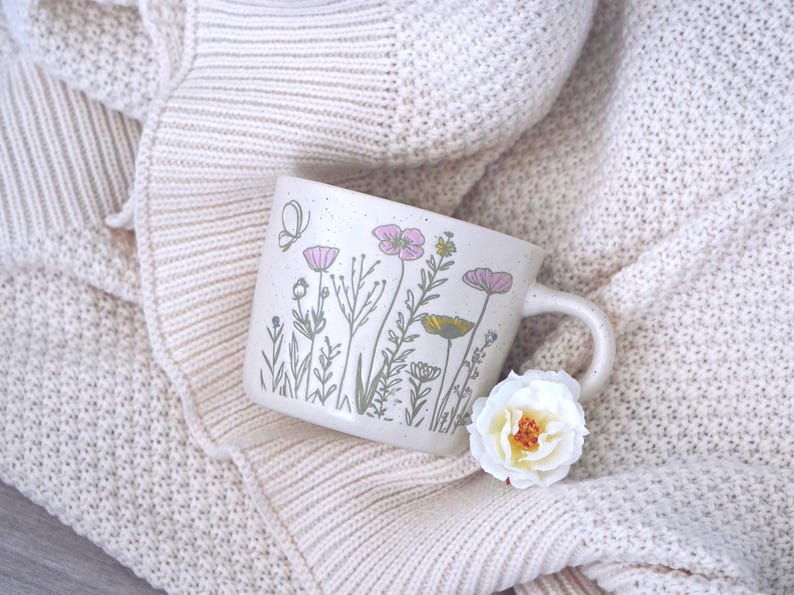 May include: A speckled off-white ceramic mug featuring a delicate botanical illustration of wildflowers in shades of pink, yellow, and green, accompanied by a small butterfly. A single white flower with a yellow center rests near the handle.