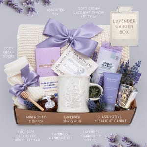 May include: A gift box filled with lavender-themed items. Includes a cream-colored knit throw (49" x 61"), a sprig mug, tea, lotion, a candle, socks, a dark chocolate bar, and a manicure kit. The box is tied with a purple bow.