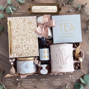 May include: A gift box labeled Quiet Reflections Box containing a linen notebook with gold line art, a gold pen, white socks with a satin ribbon, a blue tea sampler box, a lavender sprig mug, a soy candle, a linen mist spray, and honey.