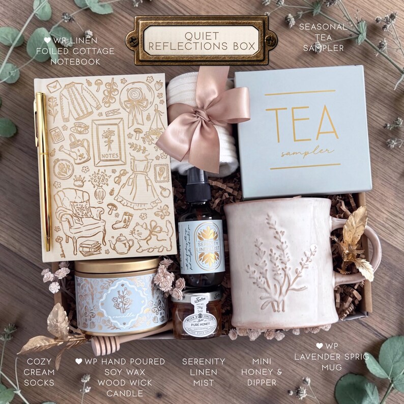 May include: A gift box labeled Quiet Reflections Box containing a linen notebook with gold line art, a gold pen, white socks with a satin ribbon, a blue tea sampler box, a lavender sprig mug, a soy candle, a linen mist spray, and honey.