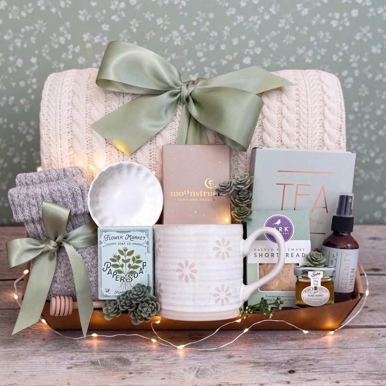 May include: A gift basket containing a cream cable-knit blanket with a sage green ribbon, a white ceramic mug with floral patterns, a box of Moonstruck chocolate, a box of tea, a bottle of spray, a jar of honey, paper soap, and gray socks.