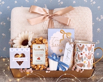 May include: A gift basket containing a cream-colored knit throw blanket with a rose gold ribbon, a bag of Popinsanity caramel popcorn, a tea sample box, a white mug with a chicken pattern and gold handle, and a small box labeled Sweet Treats.
