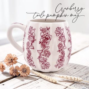 Puede incluir: Una taza de cer&aacute;mica blanca con forma de calabaza y un estampado floral tipo toile en color ar&aacute;ndano. La taza presenta un asa curva y estr&iacute;as verticales. El texto "Cranberry toile pumpkin mug" aparece escrito en negro.