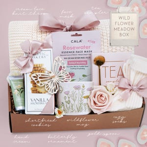 May include: A gift box filled with self-care items. Includes a cream-colored knit throw, a face mask, a wildflower mug, shortbread cookies, cozy socks, a gold spoon, and a butterfly ornament. The box is tied with a pink ribbon.