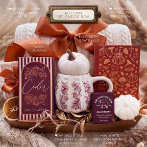 May include: An Autumn Splendor gift box containing a cream cable throw, a Spiced Apple cider box, a white pumpkin-shaped mug with cranberry floral patterns, a red notebook with gold forest motifs, a Blackberry goats milk soap, and a white pumpkin candle.