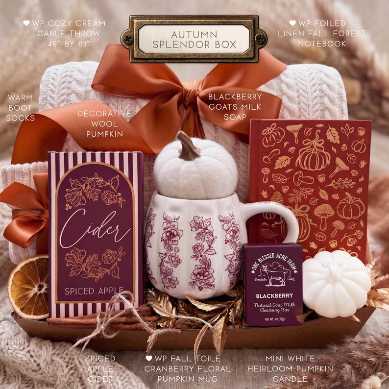 May include: An Autumn Splendor gift box containing a cream cable throw, a Spiced Apple cider box, a white pumpkin-shaped mug with cranberry floral patterns, a red notebook with gold forest motifs, a Blackberry goats milk soap, and a white pumpkin candle.