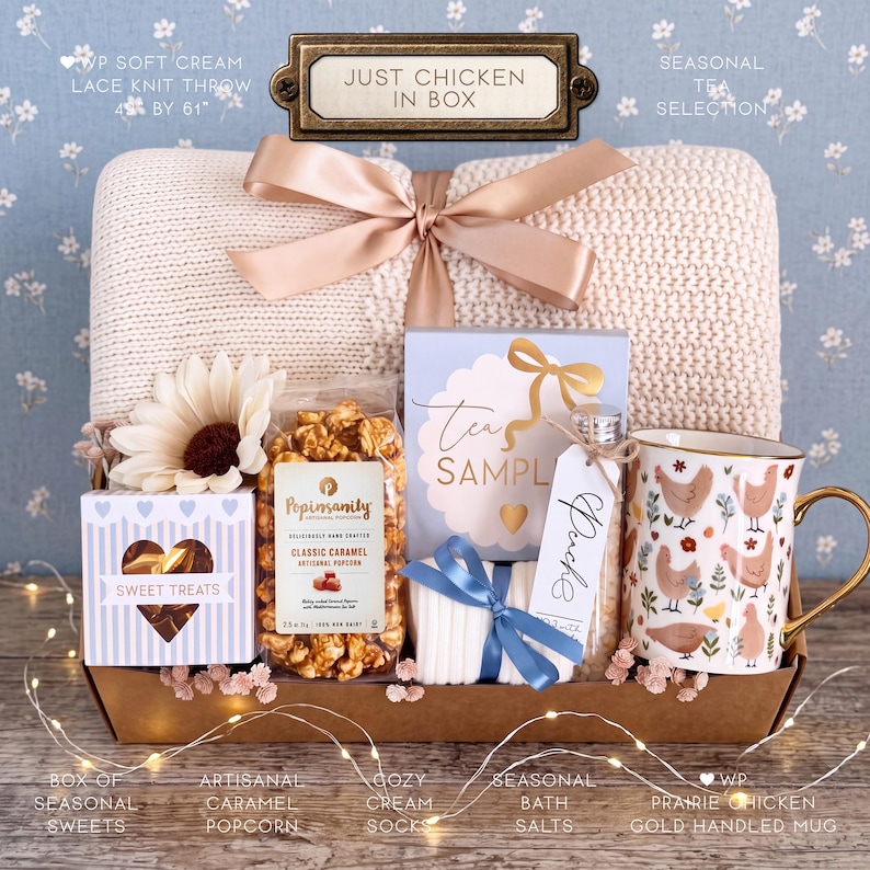 Op de afbeelding: Een geschenkmand met een cr&egrave;mekleurige gebreide plaid, een mok met kippenpatroon en gouden handvat, een zak karamelpopcorn, een doosje snoep, een theemonster, badzout en witte sokken, samengebonden met een ros&eacute;gouden lint.