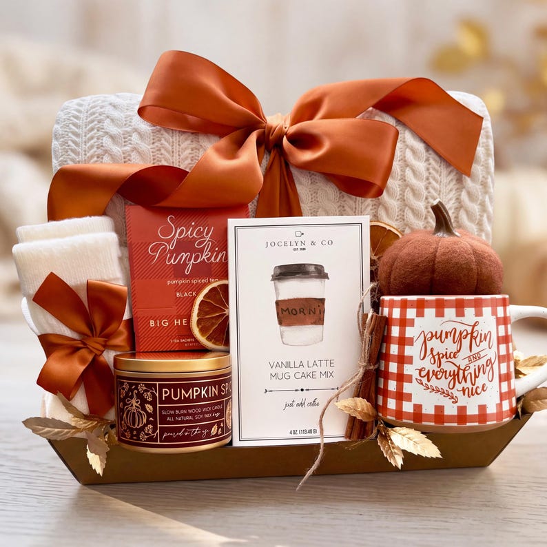 May include: A gift basket containing a white cable-knit item with a large orange ribbon, a box of Spicy Pumpkin tea, a tin of Pumpkin Spice candle, a Vanilla Latte mug cake mix, a white and orange checkered mug, and a small felt pumpkin.