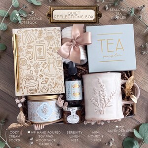 Op de afbeelding: Een geschenkdoos met het label Quiet Reflections Box bevat een linnen notitieboek met goudkleurige lijntekeningen, een gouden pen, witte sokken met een satijnen lint, een theedoos, een mok met lavendelmotief, een kaars, een spray en honing.