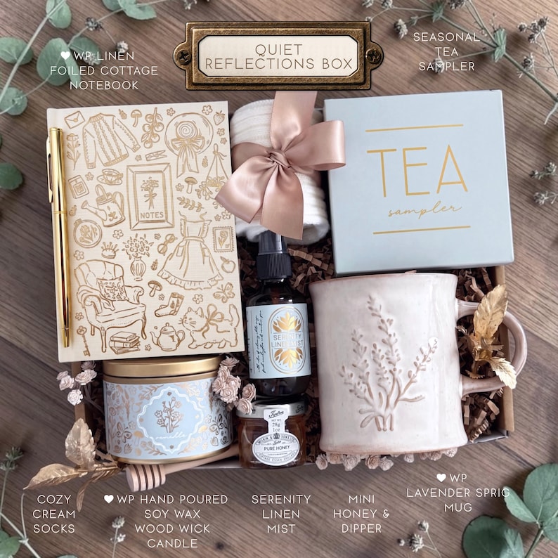 Op de afbeelding: Een geschenkdoos met het label Quiet Reflections Box bevat een linnen notitieboek met goudkleurige lijntekeningen, een gouden pen, witte sokken met een satijnen lint, een theedoos, een mok met lavendelmotief, een kaars, een spray en honing.