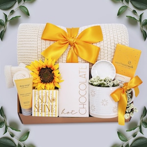 May include: A gift basket with a cream-colored knit blanket tied with a yellow satin bow. The basket includes a sunflower, body lotion, bath bomb, chocolate, a mug, and a small ceramic dish. The theme is a self-care or relaxation gift.
