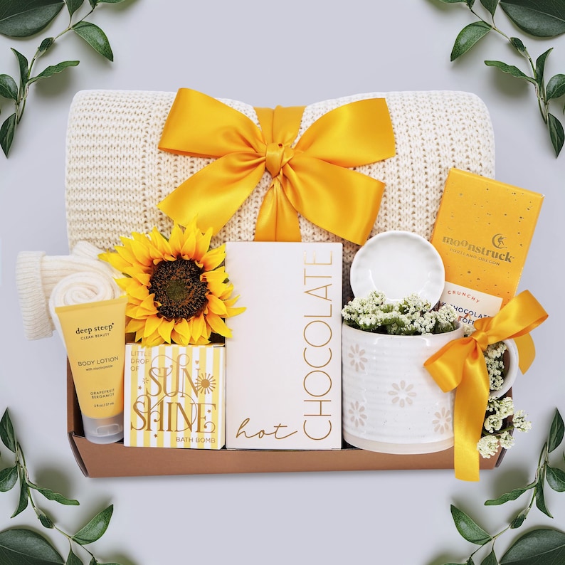 May include: A gift basket with a cream-colored knit blanket tied with a yellow satin bow. The basket includes a sunflower, body lotion, bath bomb, chocolate, a mug, and a small ceramic dish. The theme is a self-care or relaxation gift.
