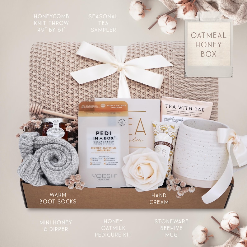 May include: A gift box with a beige honeycomb knit throw (49" x 61"), a white bow, and various self-care items. Includes a mug, socks, honey, a pedicure kit, and hand cream. The box is tied with a white ribbon.