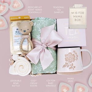 May include: A gift box containing a golden vanilla pedicure kit, a beige bear-shaped rattle, a sage green floral cotton baby swaddle tied with a pink ribbon, a seasonal tea sampler box, and a speckled ceramic mug with a floral mama design.