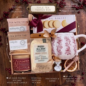 May include: A gift box containing two Tea With Tae tea packets, a box of Rustic Bakery shortbread cookies with a maroon ribbon, a Voesh Pedi in a Box kit, a white pumpkin-shaped mug with red floral patterns, honey, a dipper, and a dried orange slice.