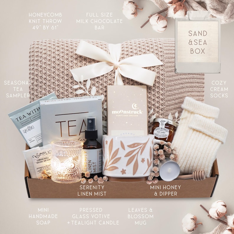 May include: A gift box filled with self-care items. Includes a honeycomb knit throw (49" x 61"), a mug with leaf design, a tea sampler, mini honey, a candle, cream socks, and other items. The box is tied with a white ribbon.