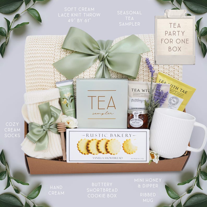 K&ouml;nnte beinhalten: Ein Geschenk-Korb mit einer weichen cremefarbenen Strickdecke (124 cm x 155 cm), Tee-Sampler, Vanille-Shortbread-Keksen, einer gerippten Tasse und anderen Tee-Party-Essentials. Der Korb ist mit einer salbeigr&uuml;nen Schleife gebunden.