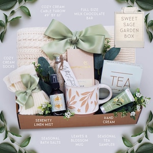 K&ouml;nnte beinhalten: Eine Geschenkbox mit einem cremefarbenen Strick&uuml;berwurf (124cm x 155cm), Socken, einer Tasse mit Bl&auml;ttermuster, Teesortiment, Handcreme, Badesalzen und einer Milchschokoladentafel. Die Box ist mit einer gr&uuml;nen Schleife und Gr&uuml;n dekoriert.